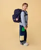 A young girl wearing a backpack and jeans stands against a plain background. Produktbild-Vorschau