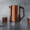 A copper electric kettle with a black handle sits on a dark countertop next to two glass mugs filled with tea. The background is a textured gray wall. Náhled obrázku produktu