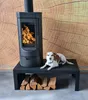 A dog is lying on a black bench in front of a black wood-burning stove with a fire inside. There are logs of wood stacked under the bench. The background is a wood-grain pattern. Náhľad obrázku produktu