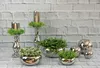 A collection of decorative silver-colored items including candle holders with greenery, planters with plants, and spherical decorative balls, arranged against a white brick wall. Produktbild-Vorschau