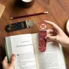 A person's hands are holding a Harry Potter themed bookmark, inserting it into an open book. Other Harry Potter themed bookmarks and a pencil are on a wooden table. Produktbild-Vorschau