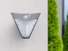 A triangular solar-powered outdoor light with a motion sensor is mounted on a textured white wall. Green bushes are visible to the right. Produktbild-Vorschau
