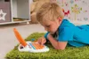 A young boy is lying on a green rug, playing with a toy laptop. Náhľad obrázku produktu