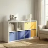 A white cube storage unit filled with books and colorful storage boxes, placed in a living room setting next to a sofa and a window with curtains. Produktbild-Vorschau