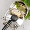 A close-up overhead view of a stainless steel pot with a glass lid, containing pasta nests, bay leaves, and a sprig of parsley. The pot is on a light gray surface with scattered peppercorns. Náhled obrázku produktu