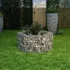 A hexagonal gabion raised garden bed filled with plants sits on a green lawn in a garden setting. Other garden elements like large planters and a wooden fence are visible in the background. Podgląd zdjęcia produktu