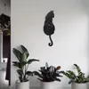 A black cat-shaped clock hangs on a white wall, with three potted plants in the foreground. Produktbild-Vorschau