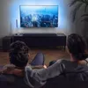 A couple is sitting on a couch watching a large TV screen, with a soundbar and subwoofer visible below the TV. Produktbild-Vorschau