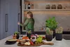 A woman is opening a cabinet in a modern kitchen, with a charcuterie board and plants on the counter. Produktbild-Vorschau