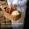 A person is holding a woven basket filled with fresh produce, including a red bell pepper and a head of cauliflower. The background shows stacks of wooden crates, suggesting a market setting. Produktbild-Vorschau
