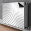 A radiator cover with a white wavy pattern is shown attached to a radiator against a concrete wall. Produktbild-Vorschau