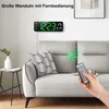 A digital wall clock is mounted on a wall above a sofa, with a hand holding a remote control pointing towards the clock. Produktbild-Vorschau