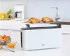 A white Braun long-slot toaster with two croissants on a warming rack, placed on a kitchen counter with a coffee maker and fruit bowl in the background. Náhled obrázku produktu