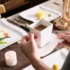 A person's hands are holding a white ceramic cup filled with coffee over a white saucer, placed on a table setting with other white ceramic plates and a lit candle. Náhled obrázku produktu