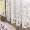 A close-up view of white sheer curtains with a gray floral pattern, draped next to a cream-colored nightstand. A fluffy rug and an open book are on the floor in the foreground. Produktbild-Vorschau