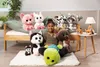 A young girl is sitting on a rug surrounded by several plush toys, with a bed in the background. Produktbild-Vorschau