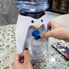 A person is using a mini penguin-shaped water dispenser to fill a glass of water. A smartphone is visible on the table. Produktbild-Vorschau