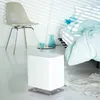 A white humidifier sits on a light-colored floor next to a bed with light blue bedding. A chair and decorative vases are in the background. Podgląd zdjęcia produktu