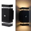 A black outdoor wall light with a motion sensor, shown both on a white background and mounted on a textured wall with light emanating from it. Produktbild-Vorschau