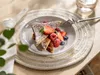 A plate of French toast with berries and powdered sugar on a placemat, with a napkin and greenery in the foreground. Produktbild-Vorschau