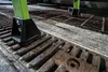 A close-up view of a green ladder leg with a black base resting on a rusty metal grate and concrete. Podgląd zdjęcia produktu