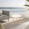A round white side table with wooden legs is placed on a rug in front of a sofa, with a view of the sea and sky through a large window. Produktbild-Vorschau