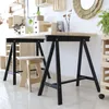 A studio shot of two black trestle legs with wooden tops, set up as a desk, with a wooden stool in front of one leg. Shelving units and decorative items are visible in the background. Podgląd zdjęcia produktu