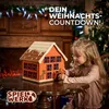 A young girl is interacting with a wooden advent calendar shaped like a house, set in a festive Christmas-themed room with a fireplace and Christmas tree. Produktbild-Vorschau