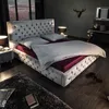 A luxurious bedroom with a large, tufted bed as the central focus. The room has dim lighting and dark walls, with a window showing a city skyline at dusk. Náhled obrázku produktu