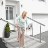 An elderly woman is carefully descending a set of outdoor stairs, using a handrail for support. She is also using two canes. The scene is in front of a modern house with a white facade and a garage door. Náhled obrázku produktu