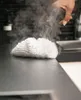 A hand holding a steam cleaner nozzle over a white cleaning pad on a dark countertop, producing steam. Produktbild-Vorschau