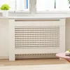 A white radiator cover with horizontal slats is shown in a room with a window and a small plant on the windowsill. Náhľad obrázku produktu
