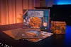 A wooden puzzle box and its packaging are displayed on a dark surface with a blurred background. Produktbild-Vorschau