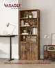 A tall, rustic-style bookshelf with two doors at the bottom, placed in a room next to a desk with a lamp and clock. A small side table with decorative items is also visible. Produktbild-Vorschau