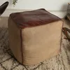 A close-up view of a cube-shaped ottoman with a brown leather top and beige fabric sides, placed on a rug. Produktbild-Vorschau