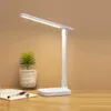 A white LED desk lamp is shown on a wooden desk next to a small potted plant and a notebook. Produktbild-Vorschau