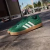 A pair of green Adidas sneakers with white stripes and a gum sole are placed on a paved surface, with a blurred background of a street scene including a bicycle. Produktbild-Vorschau