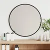 A round black-framed mirror is displayed on a white wall above a wooden shelf with various cosmetic products. Voir les images du produit