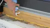 A hand is using a can of spray foam to insulate a wooden beam near a window frame. Produktbild-Vorschau