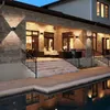A modern house with a stone facade, featuring an outdoor patio area with stairs leading down to a swimming pool. The scene is illuminated by wall lights, creating a warm ambiance. Voir les images du produit