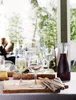 A table setting with wine glasses, a carafe of red wine, and breadsticks, with blurred people in the background. Produktbild-Vorschau