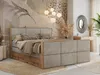 A bedroom scene featuring a large bed with a textured headboard and wooden frame, decorated with pillows and blankets. The background includes a mural, a vanity table with a mirror, and decorative plants. Produktbild-Vorschau