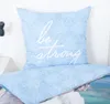 A close-up of a light blue pillow with a white floral pattern and the words "be strong" printed on it, placed on a bed. Produktbild-Vorschau