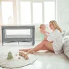A mother is playing with her baby next to a travel crib. Podgląd zdjęcia produktu