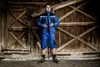 A man in workwear stands in front of a wooden barn door. Voir les images du produit