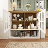A white sideboard with glass doors, filled with kitchenware and decorative items, set against a backdrop of green kitchen cabinets. Produktbild-Vorschau