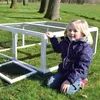 A young girl is kneeling next to a white outdoor enclosure for small animals, like rabbits or guinea pigs, in a grassy yard with trees in the background. Produktbild-Vorschau
