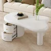 A white, uniquely shaped coffee table with two drawers, placed on a beige carpet next to a couch. Voir les images du produit