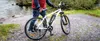 A person stands next to a white electric bicycle by a body of water. Produktbild-Vorschau
