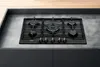 A black gas hob is installed on a grey countertop, with a wooden surface visible in the background. Voir les images du produit